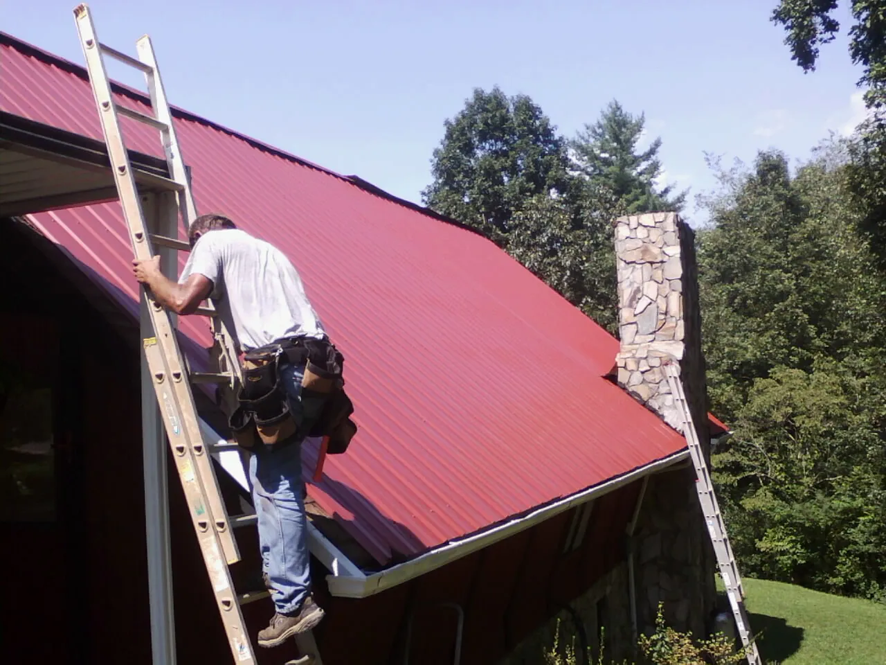 Roofer on a ladder beside a red metal roof and stone chimney — Soffit & Fascia Repair by Roofing Brothers in Winnemucca